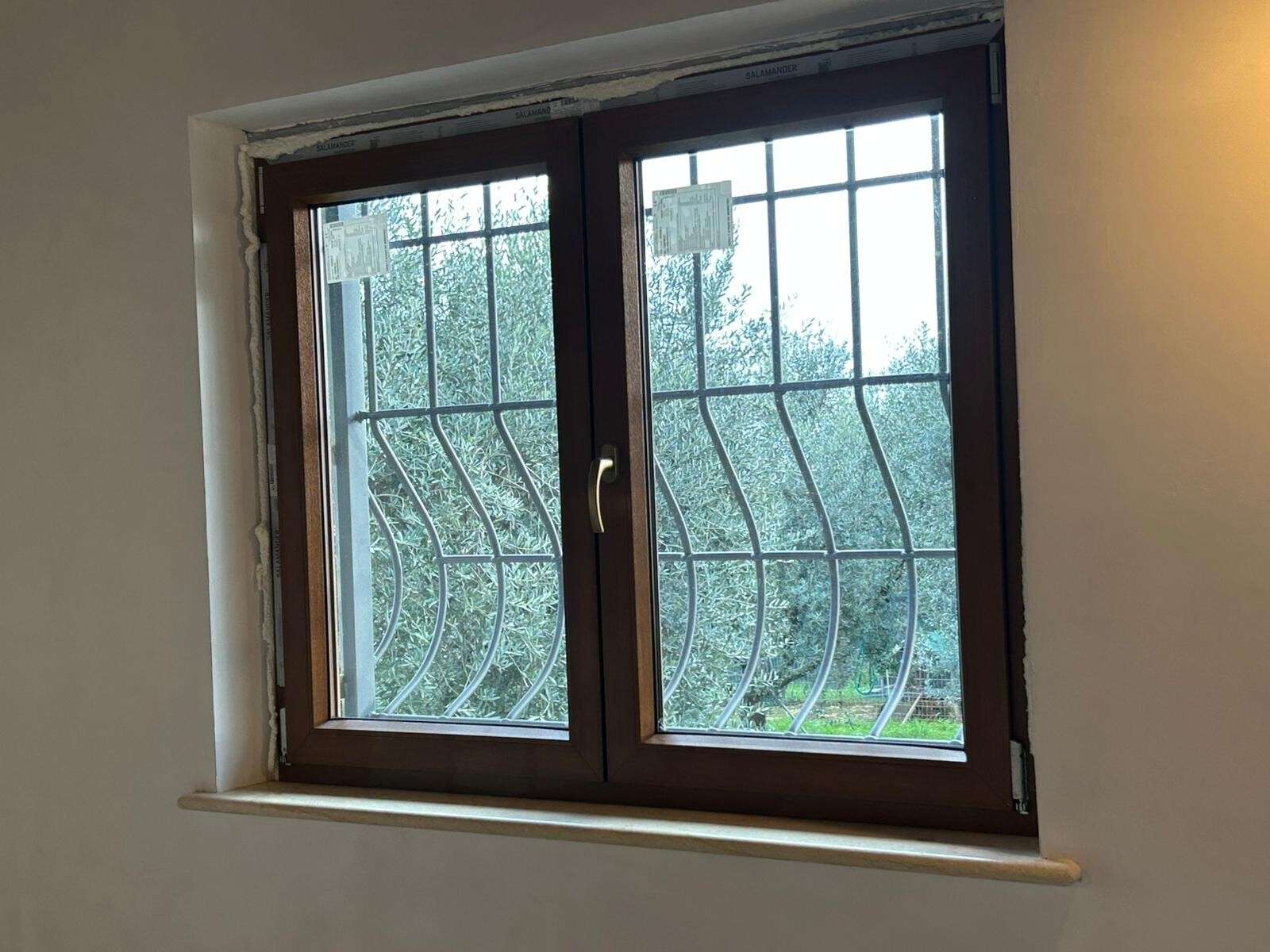 Finestra in legno scuro a due ante vista dall’interno, con grata ondulata e ulivi oltre il vetro