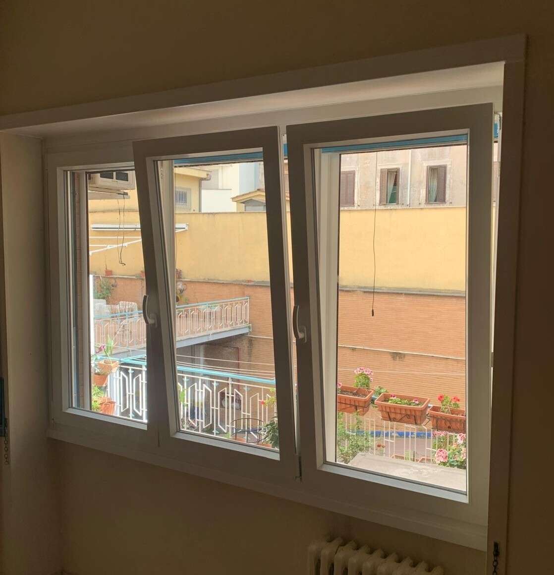 Finestra in PVC bianco a tre ante, con un’anta aperta verso un cortile con balconi